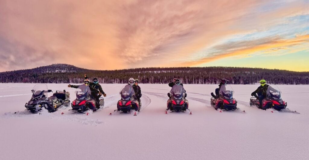 Six snowmobiles lined up on a frozen lake under a pink winter sky