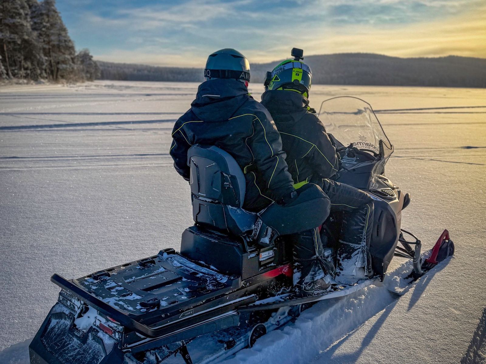 A driver and a passenger on one snowmobile, seen from behind, on a frozen lake in low sun