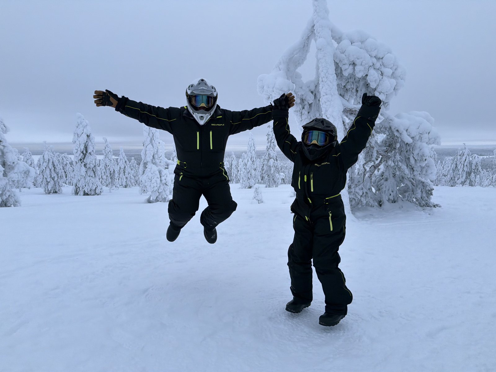 Two guests in snowmobile overalls and helmets jumping in the air on a snowy hilltop, snow-covered trees behind them
