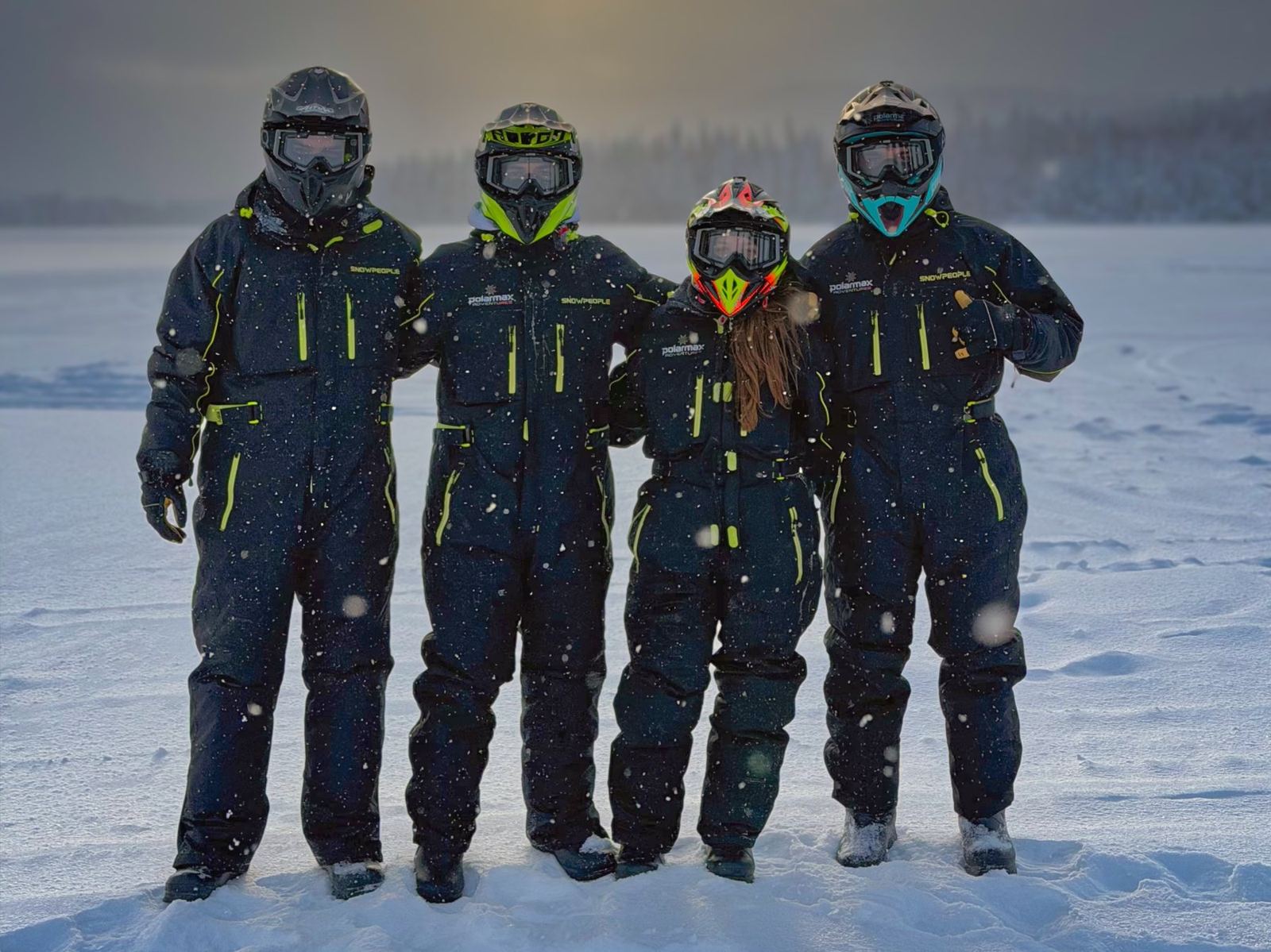 Four guests standing in a row in snowmobile overalls, helmets, goggles and boots while snow falls
