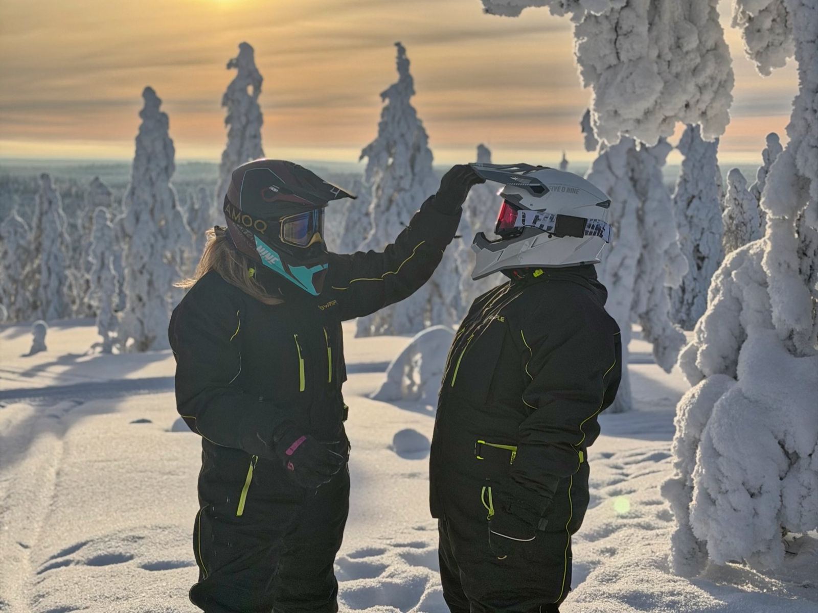 Two guests in snowmobile overalls and helmets joking around on a snowy hilltop at sunrise